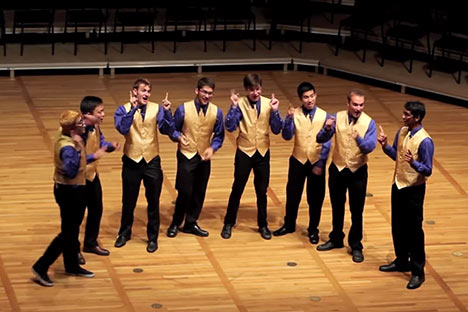 A cappela singers wearing black pants with gold vests standing together on a wood floor