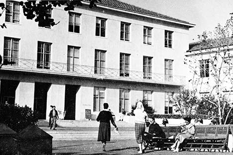 Black and white photo of building with people walking in front and two sitting on a bench