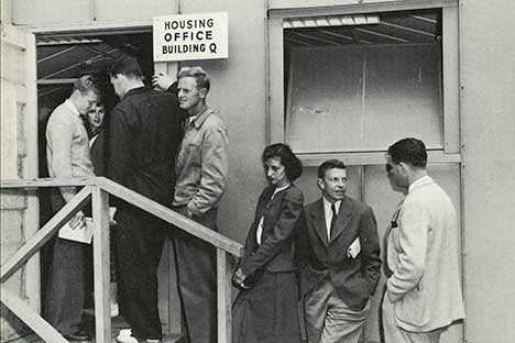 Black and white photo of people standing in line to get into a building with sign reading 'Housing Office Building Q'