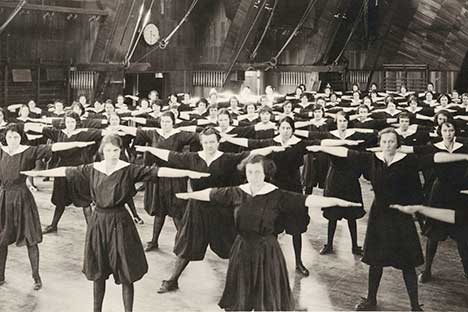 Historic black and white young women wearing dark bloomer uniforms and exercising with arms held out at shoulder level