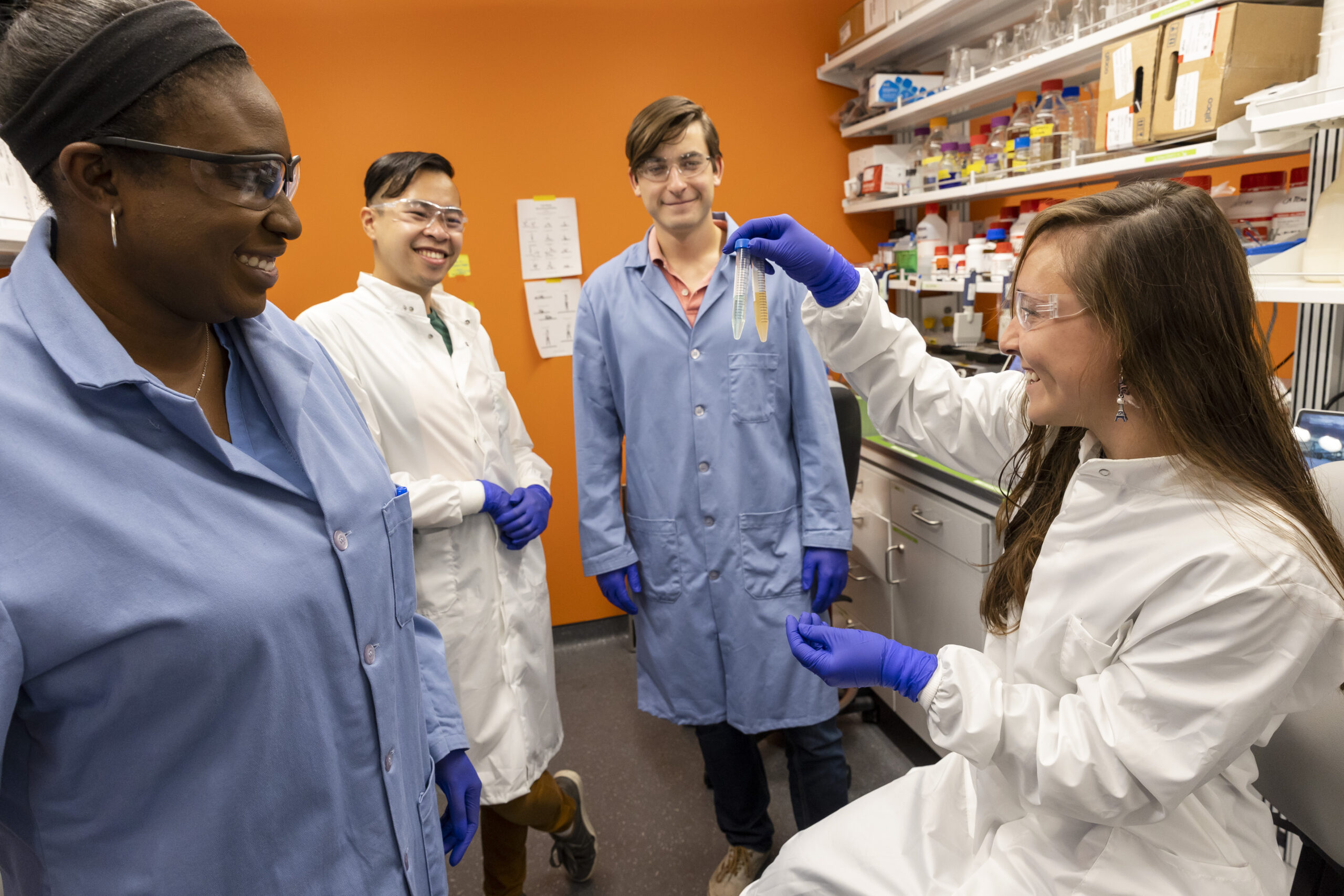 Four people are in a lab, wearing lab coats. They all look at the test tubes one woman is holding up