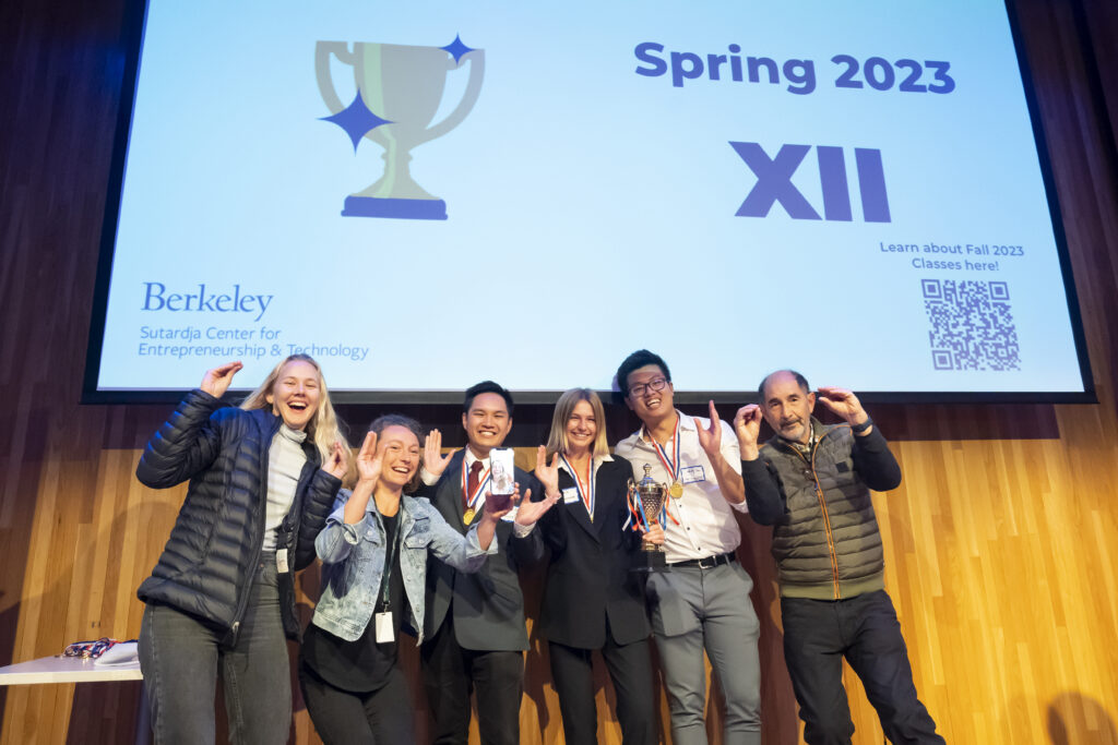 group of men and women stand with hands up, wearing medals, on stage in front of a large screen that says 