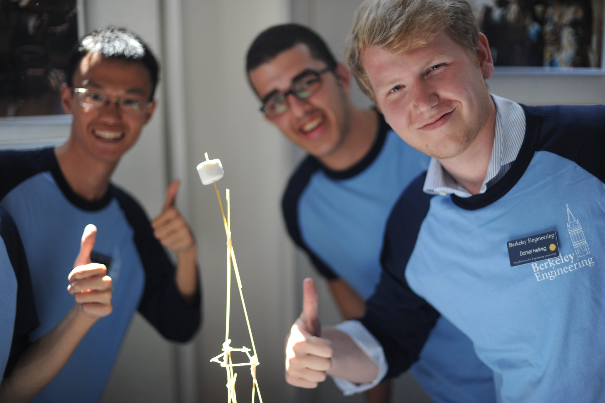 three young men in light blue shirts smile and give a thumbs up, standing next to a tower built out of spaghetti, holding up one marshmallow