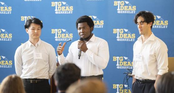 three young men stand in front of a crowd; the one in the middle speaks on a microphone