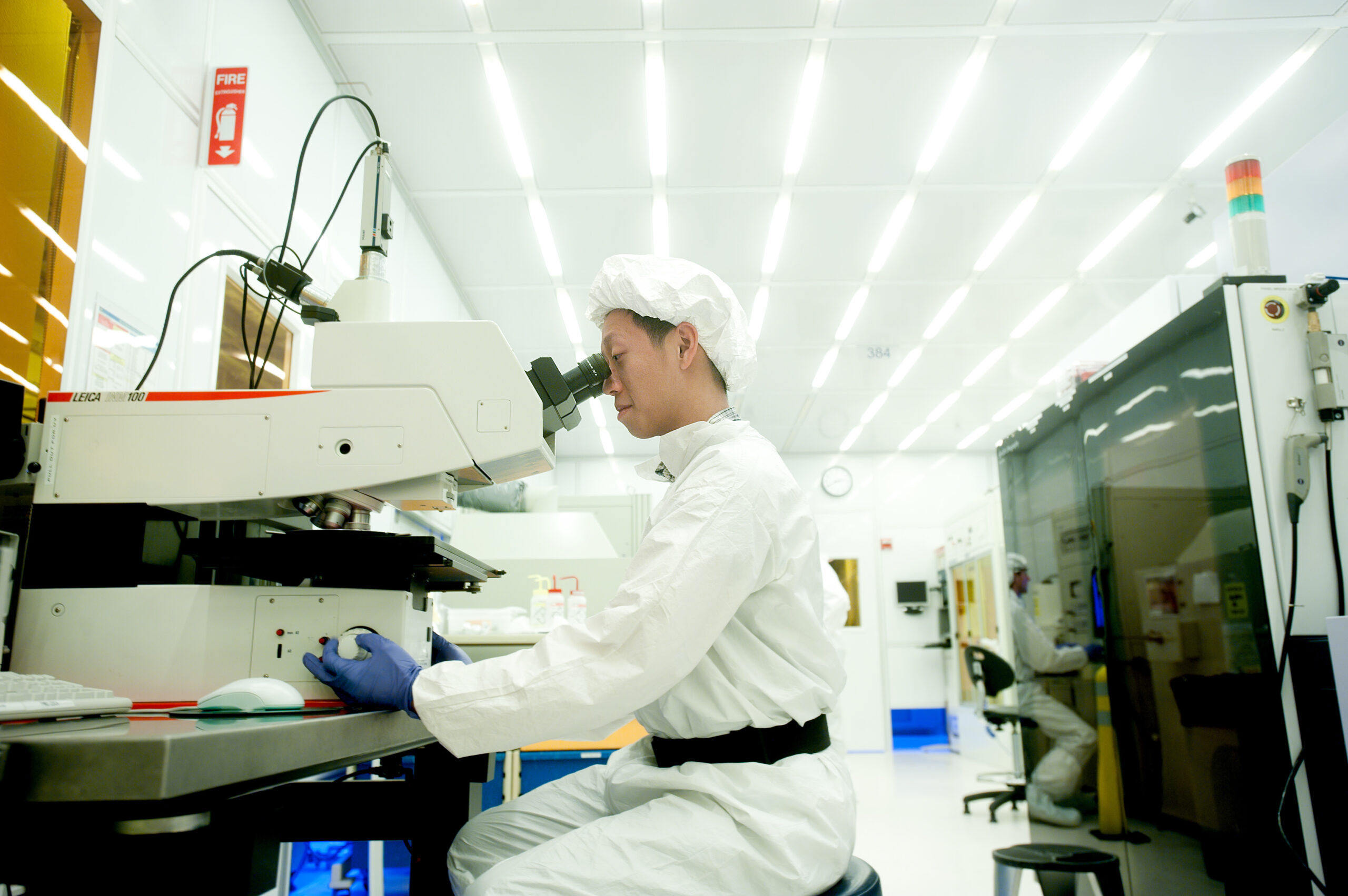 A photo of a researcher in a white lab coat and hairnet sitting at a desk looking into a large microscope.